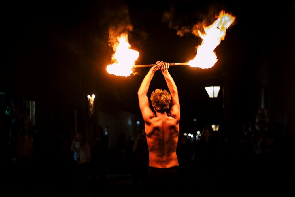 Shirtless fire performer holding a flaming staff above their head during a nighttime street performance, surrounded by a dark crowd.