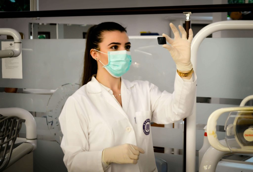 Female lab technician in a white coat and face mask holding a small black cube in a laboratory setting.