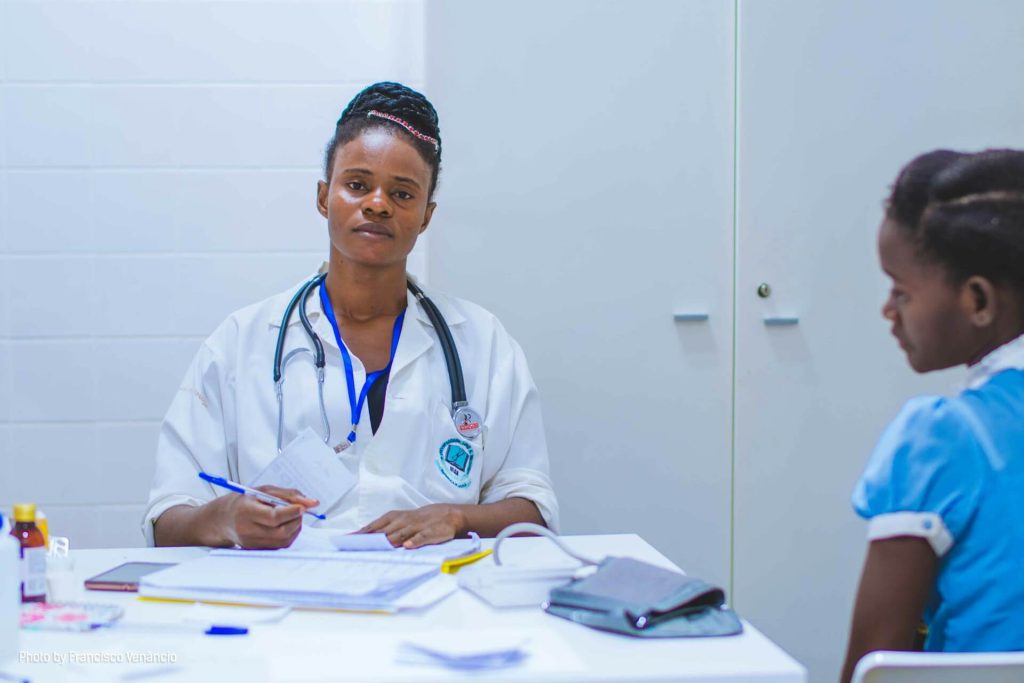 Female doctor sitting at a desk with a notepad and stethoscope, attentively listening to a young girl during a medical consultation.