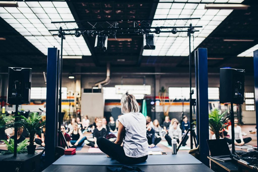 Instructor sitting cross-legged on a stage facing a large group of people in a spacious, industrial-style room during a yoga or wellness class.
