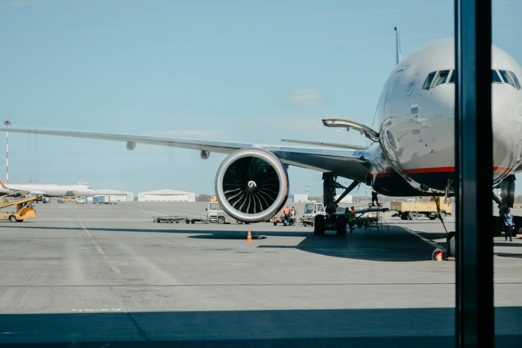 Commercial airplane parked at the gate with visible jet engine and ground crew working on the tarmac under a clear blue sky.