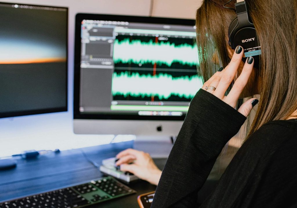 Woman wearing Sony headphones editing audio on a computer with waveforms displayed on a dual-screen setup.