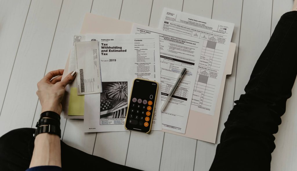 Person organizing tax documents on a white wooden floor with a smartphone calculator and pen nearby.