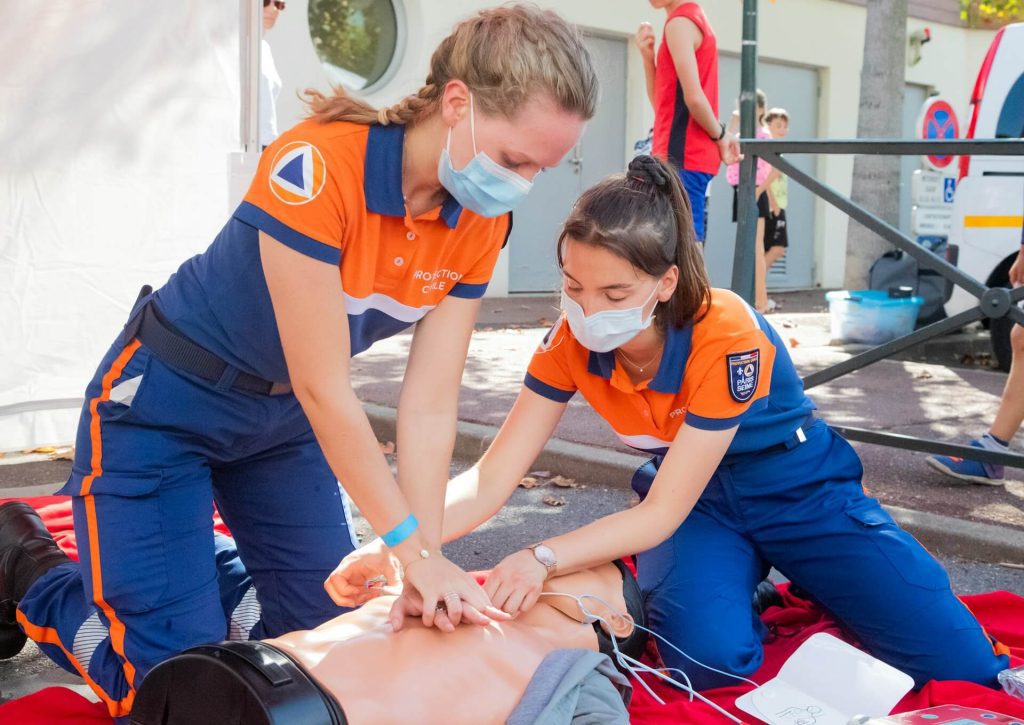 Two emergency responders in orange and blue uniforms practicing CPR on a training mannequin during a public safety demonstration.