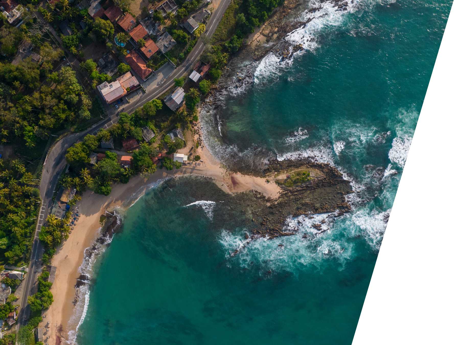 Aerial view of a rocky coastline in southern Sri Lanka with turquoise waters, sandy beach, coastal road, and houses nestled in tropical greenery.