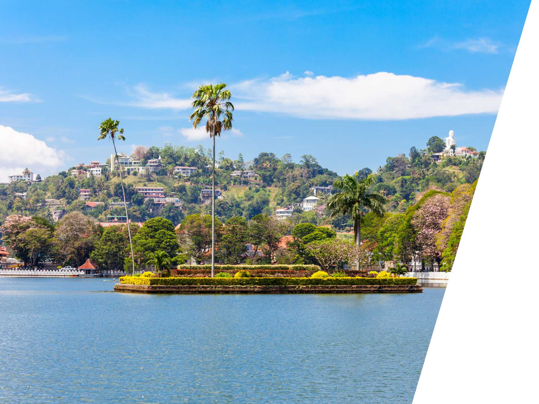 Scenic view of Kandy Lake in Sri Lanka with a small palm-filled island in the center, surrounded by hills dotted with buildings and a blue sky above.