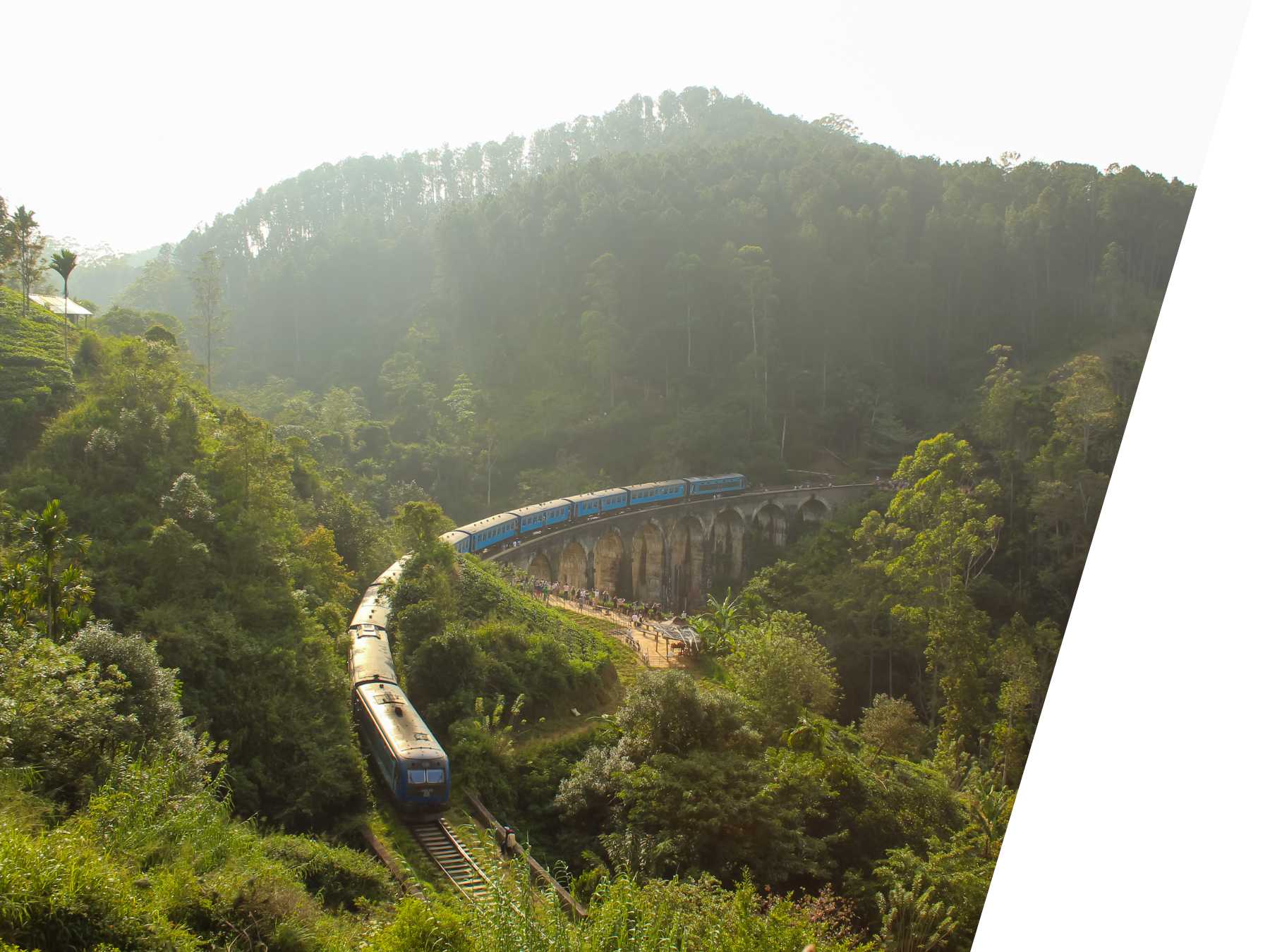 Blue train crossing the iconic Nine Arches Bridge in Ella, Sri Lanka, surrounded by lush green forest and misty hills.