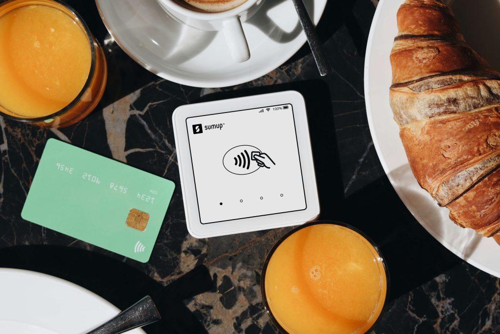 Contactless payment terminal with a green credit card, croissant, orange juice, and coffee on a dark marble table.