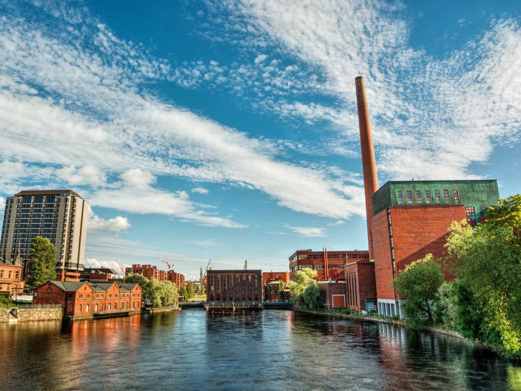 Scenic view of Tampere, Finland with the Tammerkoski river in the foreground and historic red-brick factory buildings under a vibrant blue sky with wispy clouds.