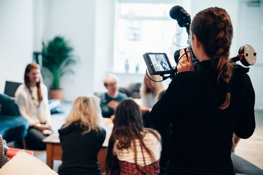 Woman filming a casual group conversation in a bright indoor setting with a professional video camera rig.