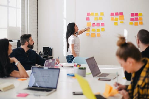 a woman is standing in front people pointing on a white board filled with yellow and pink post it notes