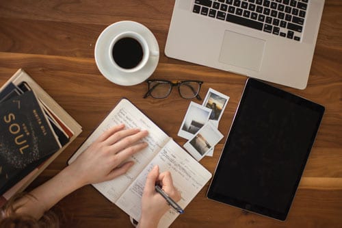 a girl is writing in a book while surrounded by a laptop, cup of coffee and photos