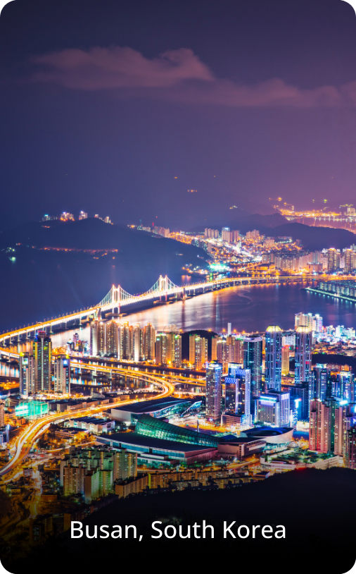 Aerial night view of Busan, South Korea, with illuminated buildings, city lights, and a dense urban landscape under a dark sky.