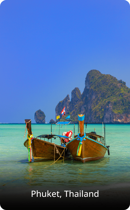 Traditional longtail boats resting on a sandy beach in Thailand, with clear water and a tropical shoreline in the background.