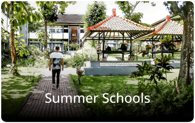 Student walking through a green campus in Bali, surrounded by lush plants, with the text “Summer Schools” overlaid on the image.